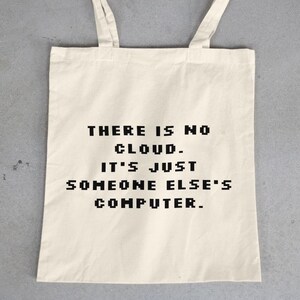 May include: Off-white tote bag with black text that reads "THERE IS NO CLOUD. IT'S JUST SOMEONE ELSE'S COMPUTER." The bag has two handles and is made of a light-colored fabric. The background is a gray concrete surface.