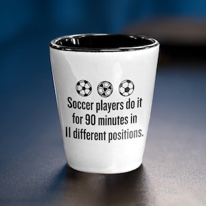 Puede incluir: Un vaso de chupito de cerámica blanca con un borde negro. Tiene un gráfico de balón de fútbol en blanco y negro y el texto "Los jugadores de fútbol lo hacen durante 90 minutos en 11 posiciones diferentes."