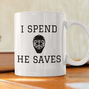May include: White ceramic coffee mug with black text that reads "I spend, he saves" and a black hockey goalie mask graphic.