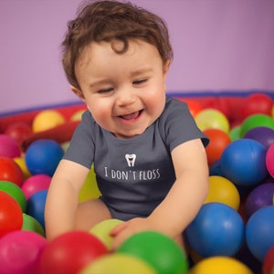 Peut inclure: Un b&eacute;b&eacute; portant un body gris avec un motif de dent blanche et le texte "Je ne passe pas de fil dentaire" est assis dans un bac &agrave; balles rempli de balles color&eacute;es.