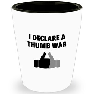May include: White shot glass with a black rim. The text "I DECLARE A THUMB WAR" is printed in bold black letters. Below the text are two thumbs up icons, one black and one gray.
