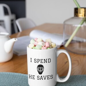 May include: White ceramic coffee mug with black text that reads "I spend, he saves" and a black hockey goalie mask graphic. The mug is filled with pastel-colored marshmallows.