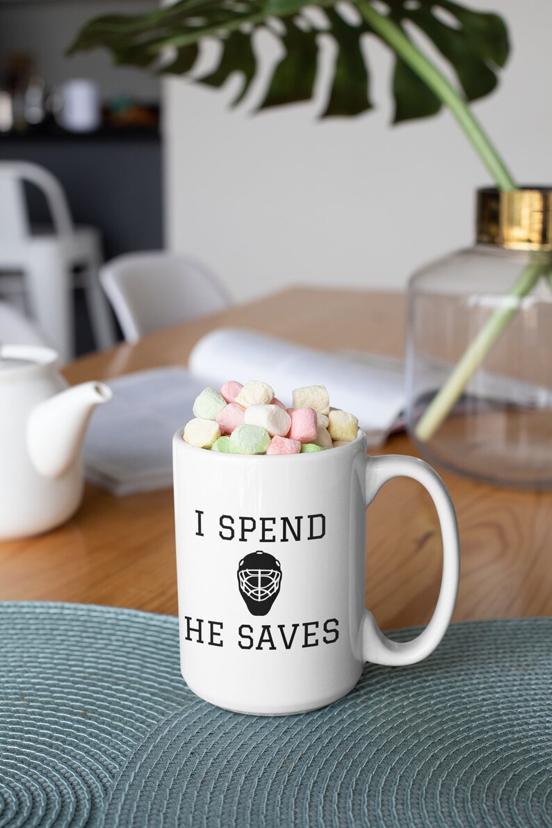 May include: White ceramic coffee mug with black text that reads "I spend, he saves" and a black hockey goalie mask graphic. The mug is filled with pastel-colored marshmallows.
