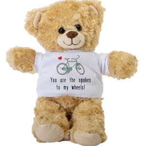Puede incluir: Un oso de peluche marrón que lleva una camiseta blanca con una bicicleta verde y un corazón rojo. La camiseta dice "You are the spokes to my wheels!"