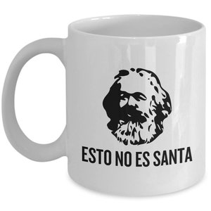 May include: White ceramic mug featuring a black silhouette of a bearded man's face. Below the image is the text "ESTO NO ES SANTA" in black capital letters. The mug has a curved handle.
