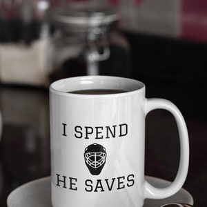 May include: White coffee mug with black text that reads "I spend he saves" and a hockey goalie mask graphic.