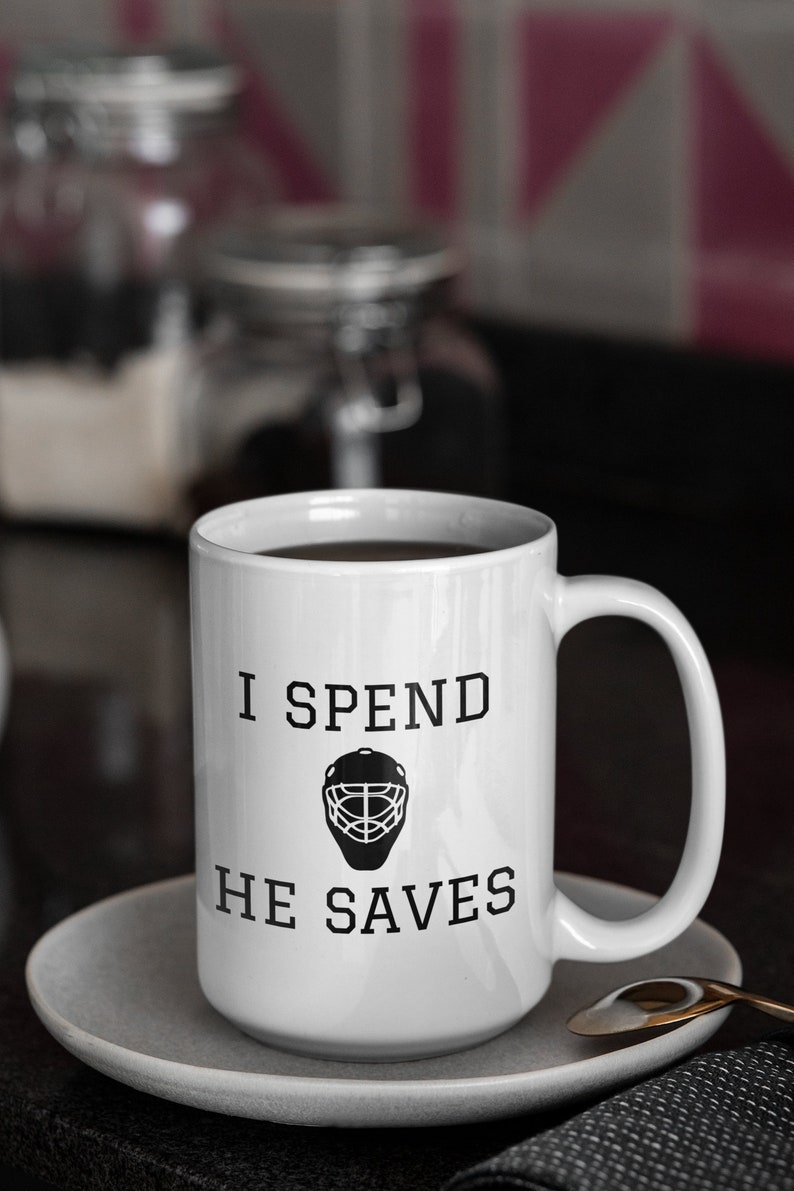 May include: White coffee mug with black text that reads "I spend he saves" and a hockey goalie mask graphic.