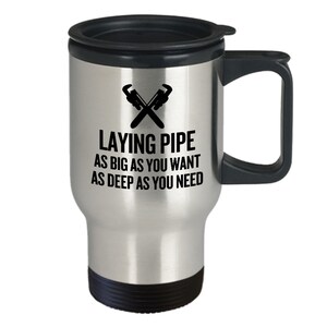 May include: A silver travel mug with a black lid and handle. The mug has a black graphic of two crossed wrenches and the text "Laying Pipe As Big As You Want As Deep As You Need".