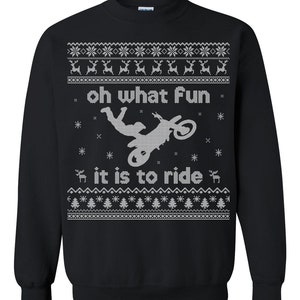Pode incluir: Camisola preta com um gráfico branco de um piloto de motocross e o texto "oh what Fun it is to ride". O design inclui flocos de neve, renas e árvores de Natal, criando um visual festivo e temático de férias.