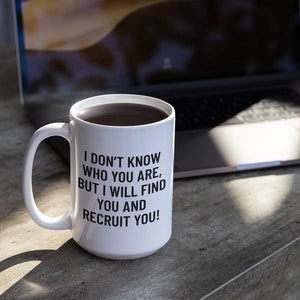 May include: White ceramic mug filled with dark liquid, featuring the text "I DON'T KNOW WHO YOU ARE, BUT I WILL FIND YOU AND RECRUIT YOU!" in black. The mug sits on a gray surface near a laptop.