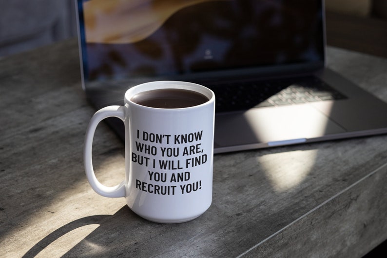 May include: White ceramic mug filled with dark liquid, featuring the text "I DON'T KNOW WHO YOU ARE, BUT I WILL FIND YOU AND RECRUIT YOU!" in black. The mug sits on a gray surface near a laptop.