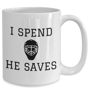 May include: White ceramic coffee mug with black text that reads "I spend, he saves" and a black silhouette of a hockey goalie mask.