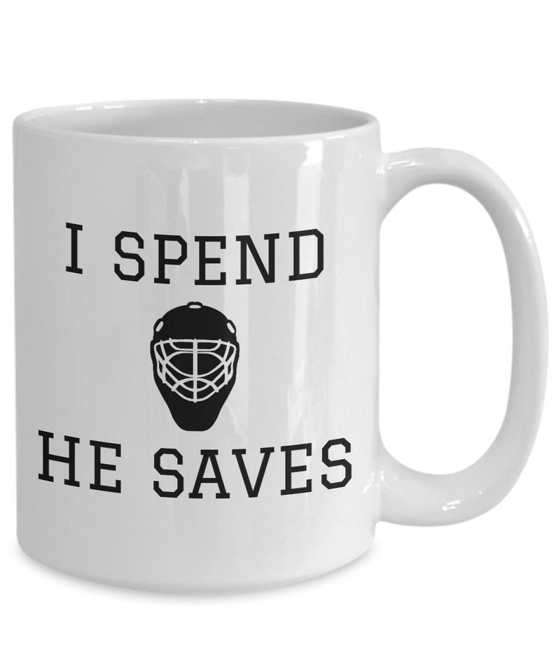 May include: White ceramic coffee mug with black text that reads "I spend, he saves" and a black silhouette of a hockey goalie mask.