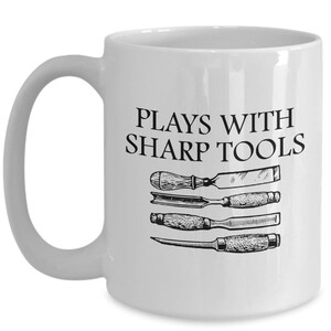 May include: White ceramic coffee mug with a black inscription that reads "Plays with sharp tools". The mug features a black and white illustration of four woodworking tools.