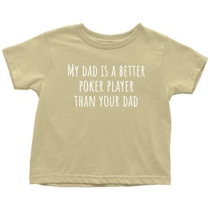 May include: A tan short-sleeved t-shirt with the text "My Dad is a Better Poker Player Than Your Dad" printed in white.
