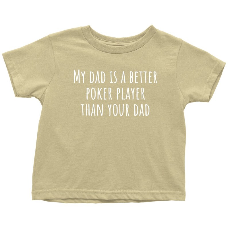 May include: A tan short-sleeved t-shirt with the text "My Dad is a Better Poker Player Than Your Dad" printed in white.