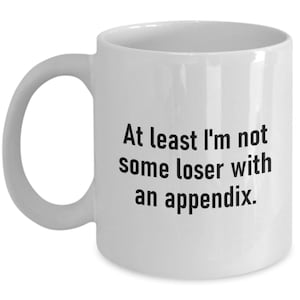 Könnte beinhalten: Weiße Keramik-Kaffeetasse mit schwarzem Text, der lautet: "Zumindest bin ich kein Loser mit einem Blinddarm."