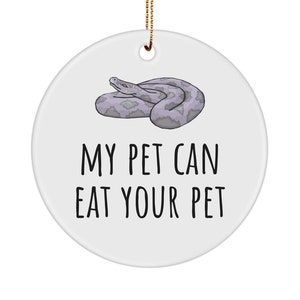 Puede incluir: Adorno redondo de cerámica blanca con una cuerda dorada para colgar. Presenta una ilustración de dibujos animados de una serpiente gris y el texto "MY PET CAN EAT YOUR PET" en negro.