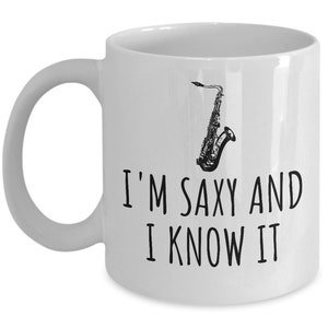 Puede incluir: Taza de café de cerámica blanca con una ilustración en blanco y negro de un saxofón y el texto "I'm saxy and I know it".