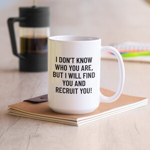 May include: White ceramic mug with a black handle and the text "I DON'T KNOW WHO YOU ARE, BUT I WILL FIND YOU AND RECRUIT YOU!". A French press and a notebook are in the background.