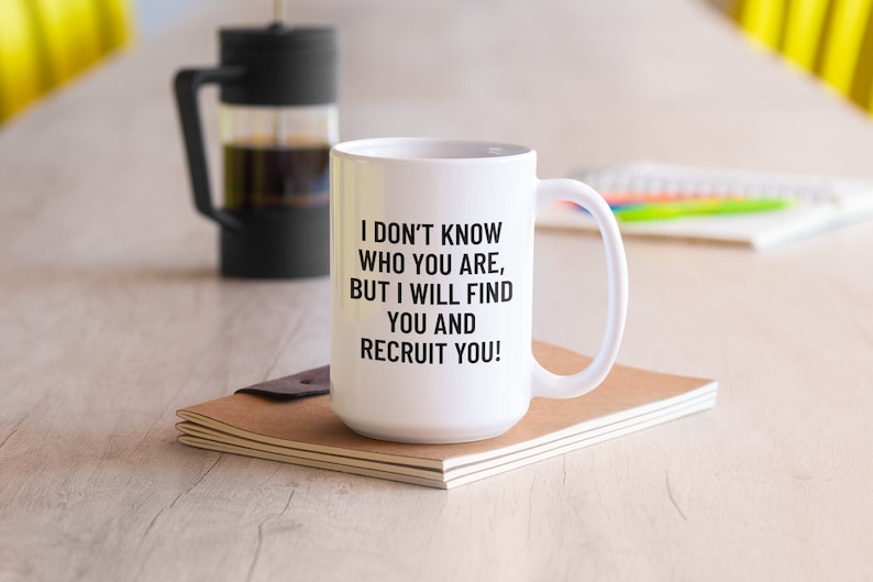 May include: White ceramic mug with a black handle and the text "I DON'T KNOW WHO YOU ARE, BUT I WILL FIND YOU AND RECRUIT YOU!". A French press and a notebook are in the background.
