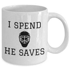 May include: White ceramic coffee mug with black text that reads "I spend, he saves" and a black silhouette of a hockey goalie mask.
