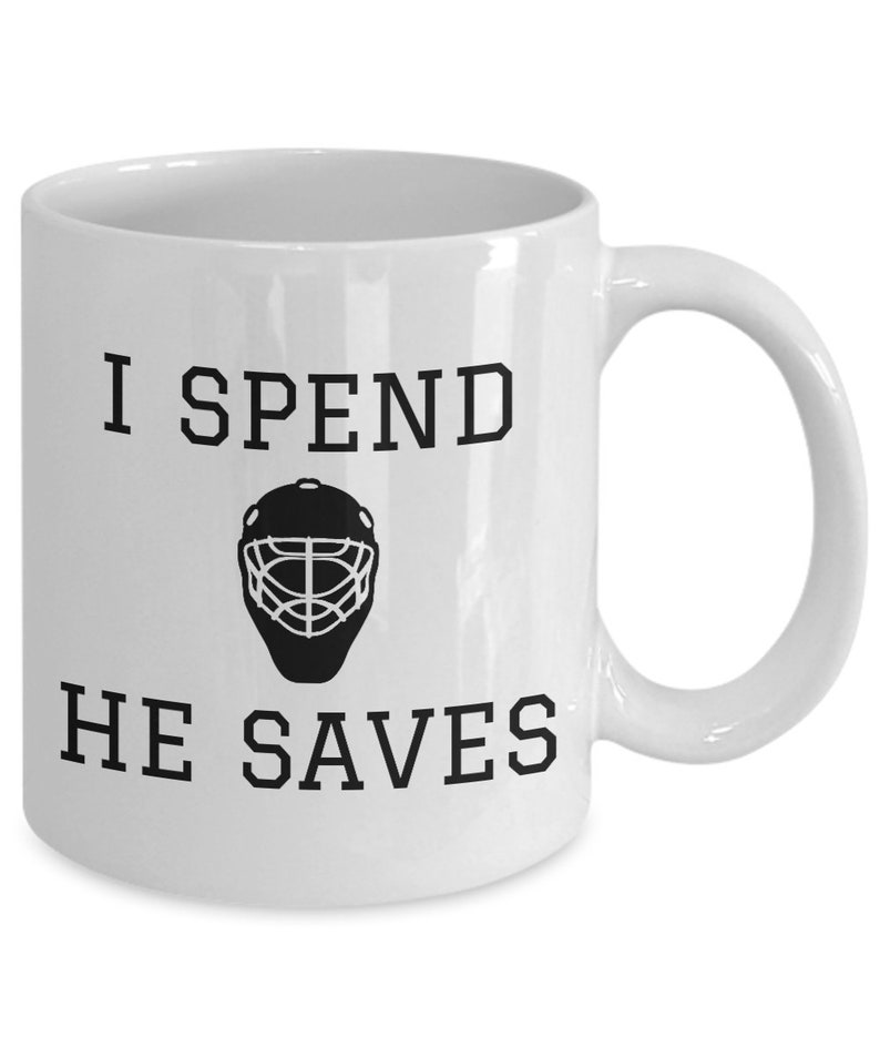 May include: White ceramic coffee mug with black text that reads "I spend, he saves" and a black silhouette of a hockey goalie mask.