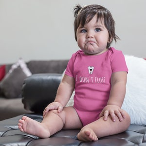 Peut inclure: Un body b&eacute;b&eacute; rose avec une dent de dessin anim&eacute; blanche et le texte "Je ne passe pas de fil dentaire". Un b&eacute;b&eacute; porte le body et est assis sur un canap&eacute; en cuir noir.