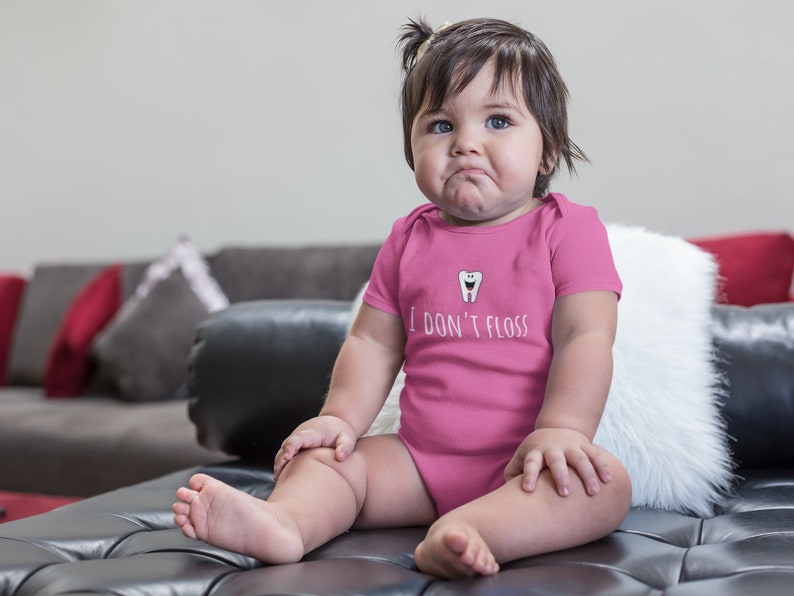 Peut inclure: Un body b&eacute;b&eacute; rose avec une dent de dessin anim&eacute; blanche et le texte "Je ne passe pas de fil dentaire". Un b&eacute;b&eacute; porte le body et est assis sur un canap&eacute; en cuir noir.