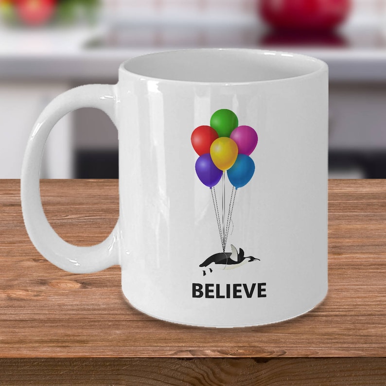 May include: White ceramic mug with a penguin holding onto colorful balloons that say "BELIEVE".