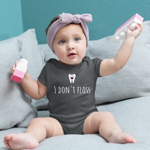 Peut inclure: Un b&eacute;b&eacute; portant un body gris fonc&eacute; avec une dent de dessin anim&eacute; blanche et le texte "I don't floss". Le b&eacute;b&eacute; est assis sur un canap&eacute; gris et tient un jouet rose et blanc.