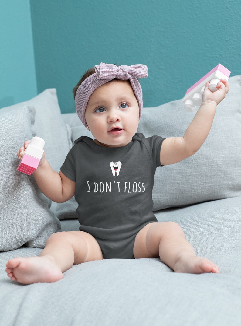 Peut inclure: Un b&eacute;b&eacute; portant un body gris fonc&eacute; avec une dent de dessin anim&eacute; blanche et le texte "I don't floss". Le b&eacute;b&eacute; est assis sur un canap&eacute; gris et tient un jouet rose et blanc.