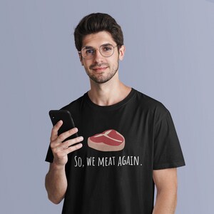 May include: A black t-shirt with a white graphic of a steak and the text "So, we meat again."