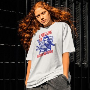 May include: White t-shirt featuring a blue and red graphic of a bird reading a newspaper. The text on the shirt says "I FEEL LIKE I'M BEING WATCHED". The t-shirt has a relaxed, oversized style.