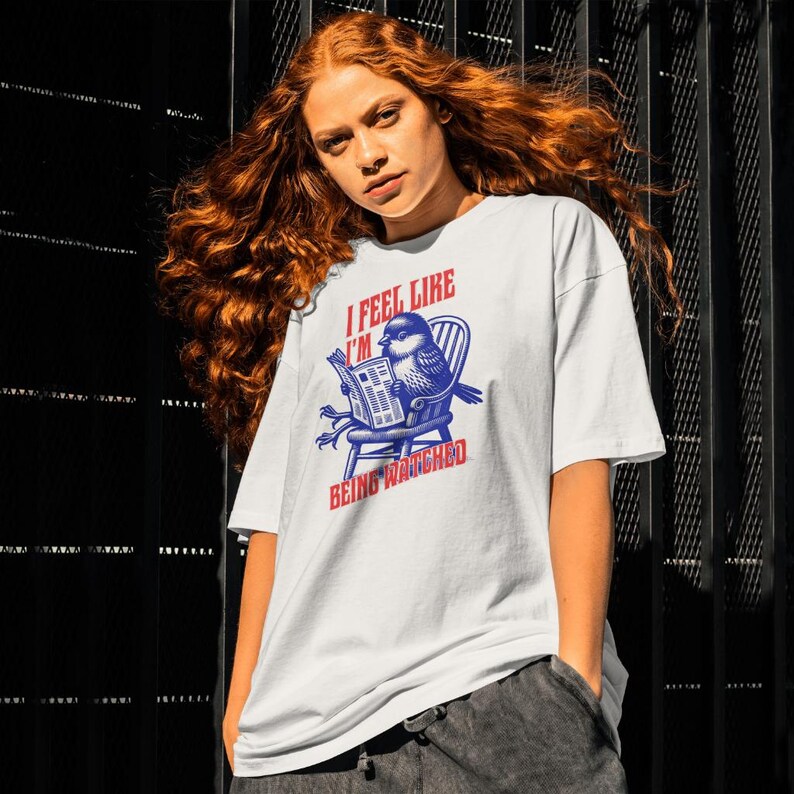 May include: White t-shirt featuring a blue and red graphic of a bird reading a newspaper. The text on the shirt says "I FEEL LIKE I'M BEING WATCHED". The t-shirt has a relaxed, oversized style.