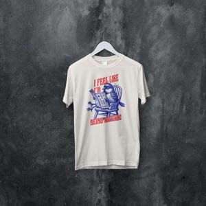 May include: White t-shirt featuring a blue bird reading a newspaper, seated in a chair. The text on the shirt reads "I FEEL LIKE I'M BEING WATCHED". The design is in blue and red.