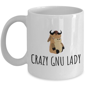 May include: White ceramic mug featuring a cartoon gnu illustration and the text "CRAZY GNU LADY". The gnu is brown and black with a serious expression. The mug has a curved handle, perfect for tea or coffee.