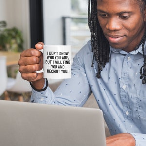 May include: A white ceramic mug with the text "I DON'T KNOW WHO YOU ARE, BUT I WILL FIND YOU AND RECRUIT YOU!" in black. The mug is held by a person in a blue button-down shirt, working on a laptop.