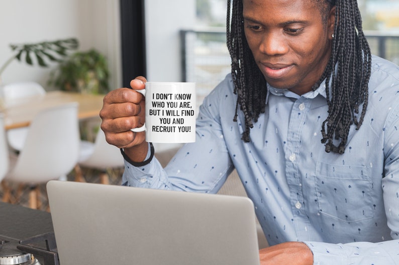 May include: A white ceramic mug with the text "I DON'T KNOW WHO YOU ARE, BUT I WILL FIND YOU AND RECRUIT YOU!" in black. The mug is held by a person in a blue button-down shirt, working on a laptop.