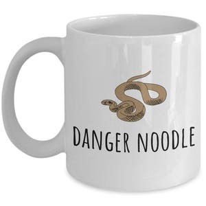 Peut inclure: Une tasse à café en céramique blanche avec une illustration de serpent brun et le texte "DANGER NOODLE" imprimé en noir.
