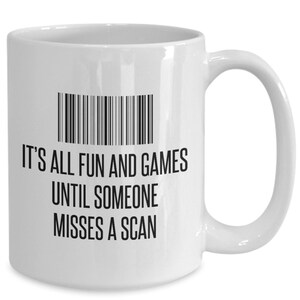May include: White ceramic mug with a black barcode design and the text "IT'S ALL FUN AND GAMES UNTIL SOMEONE MISSES A SCAN". The mug has a curved handle and a glossy finish.