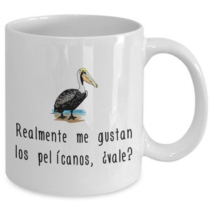 May include: White ceramic mug with a pelican illustration. The pelican has black and brown feathers, a yellow head, and a long beak. The mug has the text "Realmente me gustan los pelícanos, ¿vale?" in black.