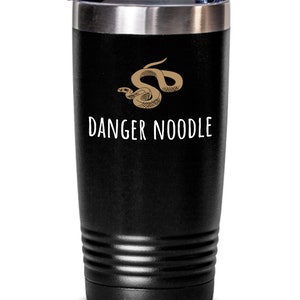 Può includere: Bicchiere da viaggio nero con bordo argentato e coperchio trasparente. Il bicchiere presenta un'illustrazione marrone di un serpente e le parole "DANGER NOODLE" in bianco, un design umoristico.