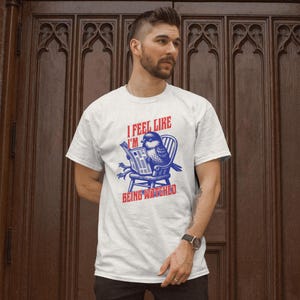 May include: White t-shirt featuring a blue and red graphic of a bird reading a newspaper. The text on the shirt says "I FEEL LIKE I'M BEING WATCHED".