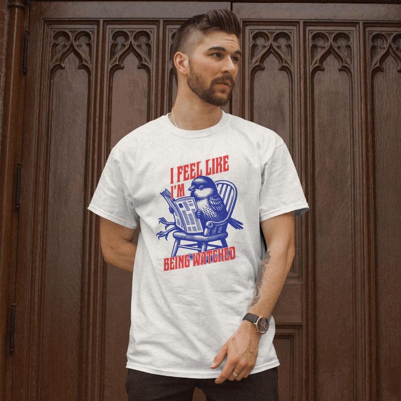May include: White t-shirt featuring a blue and red graphic of a bird reading a newspaper. The text on the shirt says "I FEEL LIKE I'M BEING WATCHED".
