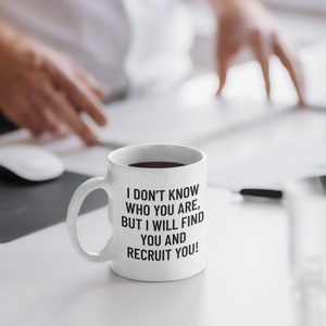 May include: White ceramic mug with the text "I DON'T KNOW WHO YOU ARE, BUT I WILL FIND YOU AND RECRUIT YOU!" in black. The mug is filled with a dark liquid, likely coffee or tea. The mug is on a white desk.