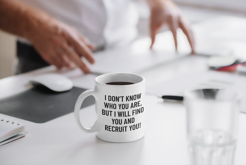 May include: White ceramic mug with the text "I DON'T KNOW WHO YOU ARE, BUT I WILL FIND YOU AND RECRUIT YOU!" in black. The mug is filled with a dark liquid, likely coffee or tea. The mug is on a white desk.