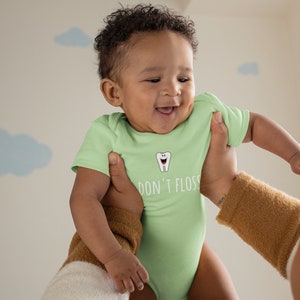 Peut inclure: Un b&eacute;b&eacute; portant une grenouill&egrave;re vert clair avec une dent de dessin anim&eacute; blanche et le texte "Don't Floss" imprim&eacute; dessus. Le b&eacute;b&eacute; est tenu par une personne.