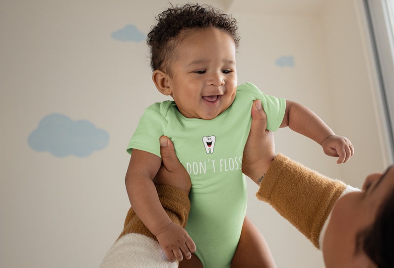 Peut inclure: Un b&eacute;b&eacute; portant une grenouill&egrave;re vert clair avec une dent de dessin anim&eacute; blanche et le texte "Don't Floss" imprim&eacute; dessus. Le b&eacute;b&eacute; est tenu par une personne.
