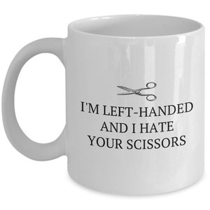 May include: White ceramic coffee mug with a black graphic of a pair of scissors and the text "I'm left-handed and I hate your scissors".